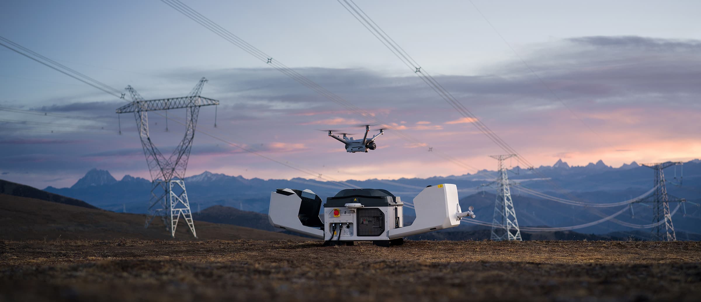 En DJI Matrice 4D-drönare lyfter från en DJI Dock 3-drönarstation i ett öppet landskap med kraftledningar och berg i bakgrunden vid solnedgång.