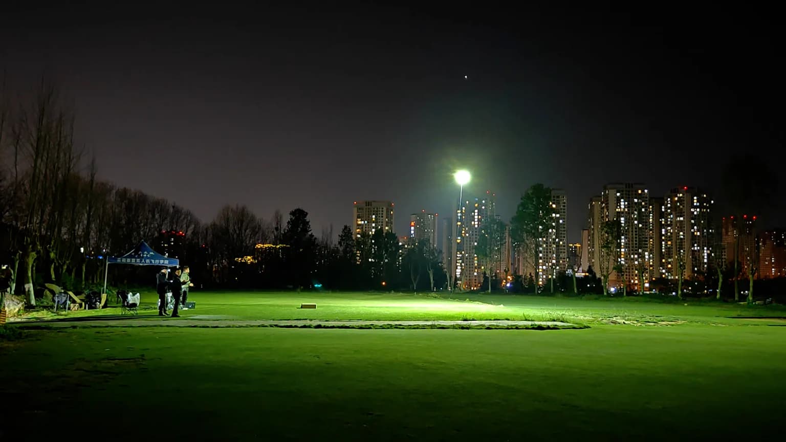 En nattlig scen med upplyst av CZ100 Tethered Hover Light och en stadssilhuett i bakgrunden, där människor samlas under ett tält.