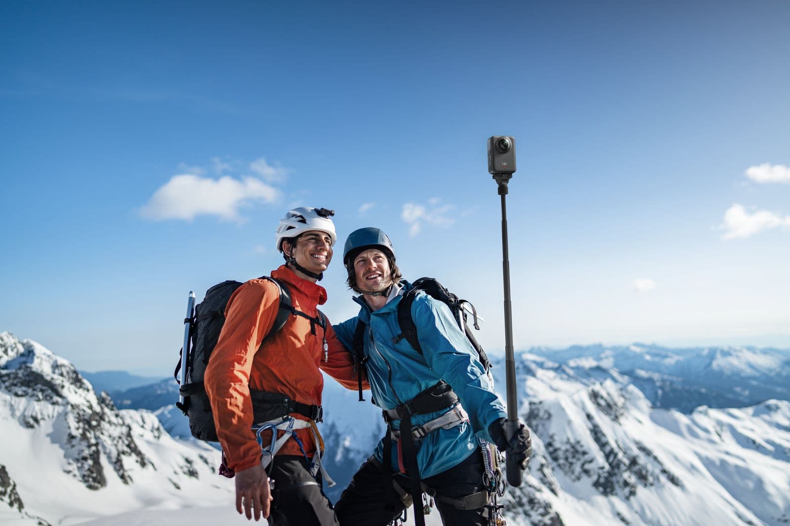 Två klättrare i färgglada jackor poserar för en selfie med en kamera på en stav, omgivna av snötäckta bergstoppar under en klarblå himmel.